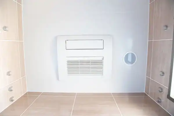 A white wall-mounted air vent on a tiled bathroom wall, surrounded by light brown tiles and a round pipe opening to the right.