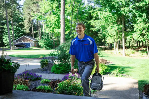 A man in a blue polo shirt and gray pants walks toward a house carrying a small gray bag, surrounded by lush greenery and a garden in the background.